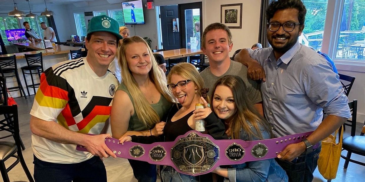 Group of friends proudly holding a purple trivia champion belt, smiling inside a cozy bar.